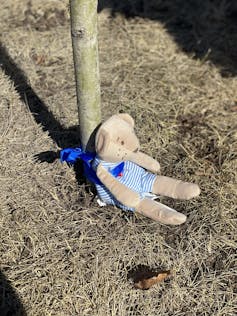 a stuffed teddy bear sits beside a wooden marker in the ground.