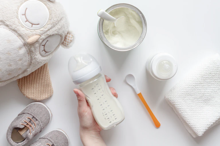 A hand holds a baby bottle containing milk. Scattered around are various baby artefacts.