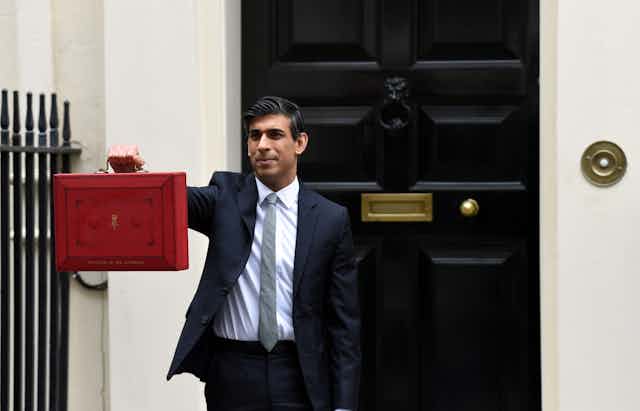 A man in a suit stands outside a black door holding a red briefcase