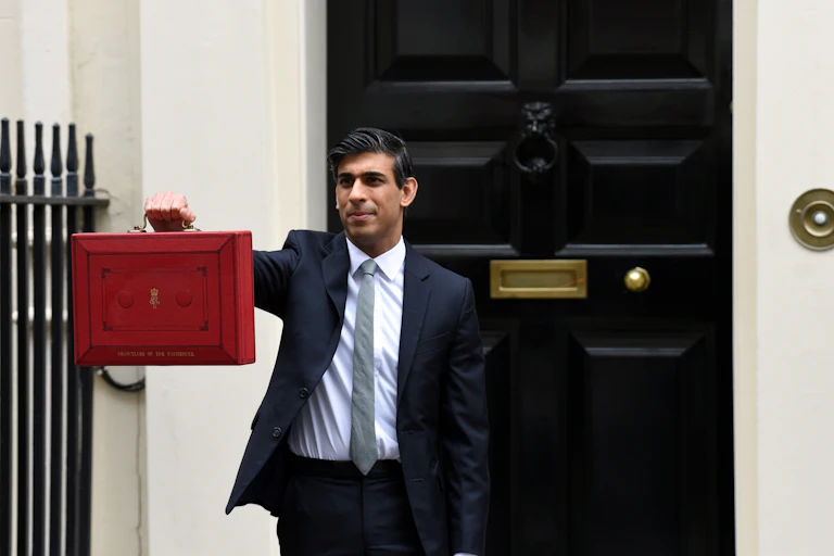 A man in a suit stands outside a black door holding a red briefcase