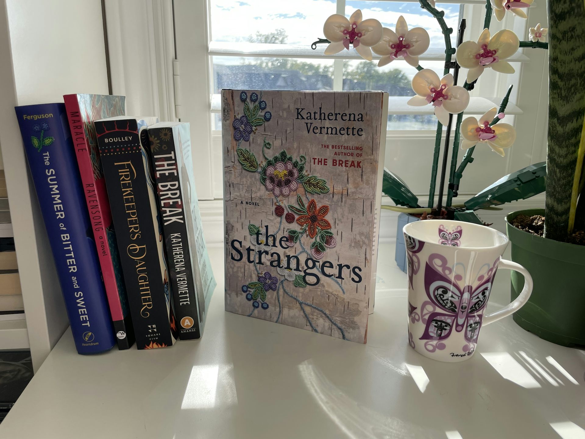 Books seen by a windowsill next to a mug with a butterfly on it.