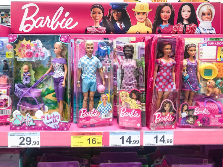 A Ken doll wearing blue is in a Barbie box on a shelf, surrounded by other Barbie dolls.