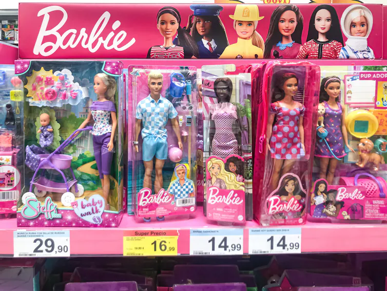 A Ken doll wearing blue is in a Barbie box on a shelf, surrounded by other Barbie dolls.