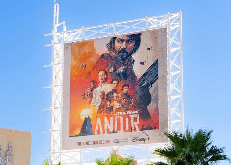 A billboard against a blue sky shows a man with dark hair and a beard in front of other people, with the words
