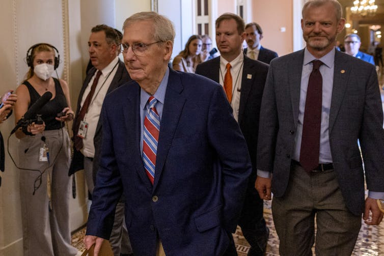 A gray-haired man in a blue blazer walks in a building followed by several other people.