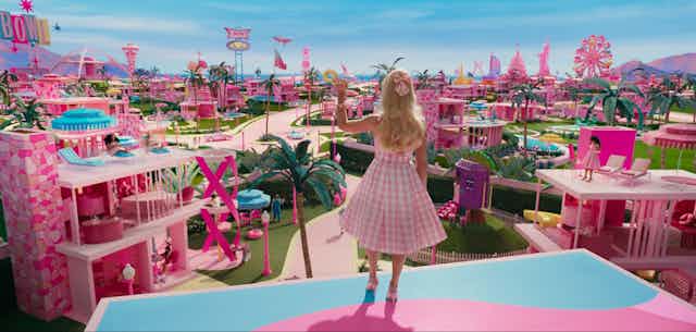 The back of a blonde woman wearing a pink dress is seen looking over a pink series of buildings with palm trees and a blue sky. 