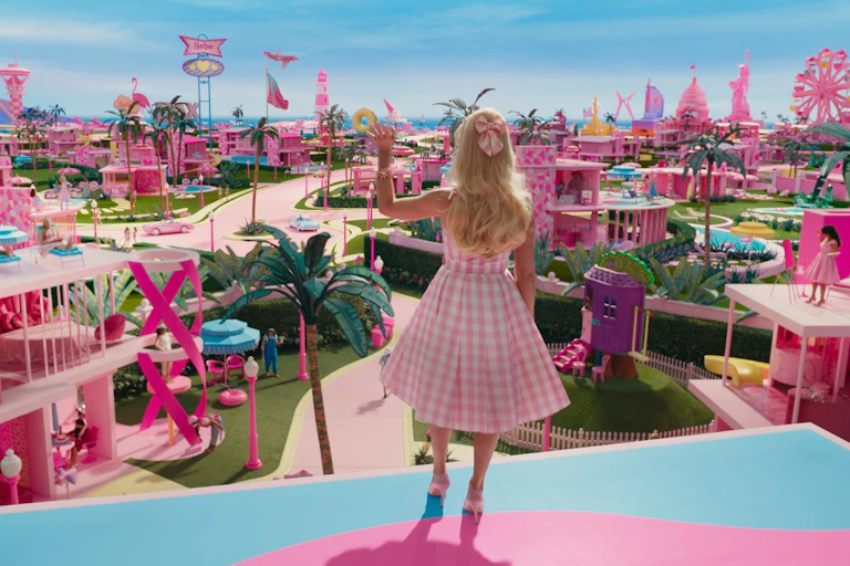 The back of a blonde woman wearing a pink dress is seen looking over a pink series of buildings with palm trees and a blue sky.