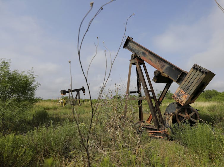 Photo of a rusted oil pump in an overgrown field in Texas. Rusted parts are piled beside it.