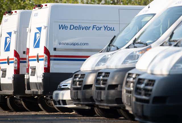 U.S. Postal Service vehicles of different sizes are parked in a lot, some facing in, some facing out. The side of one reads: 'We deliver for you'