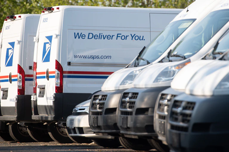 U.S. Postal Service vehicles of different sizes are parked in a lot, some facing in, some facing out. The side of one reads: 'We deliver for you'