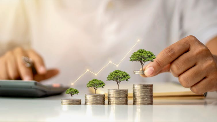 Four stacks of coins sit on a table, each increasing in size. A small tree sits on three of the stacks and a hand is placing a fourth tree on the last, tallest stack of coins.