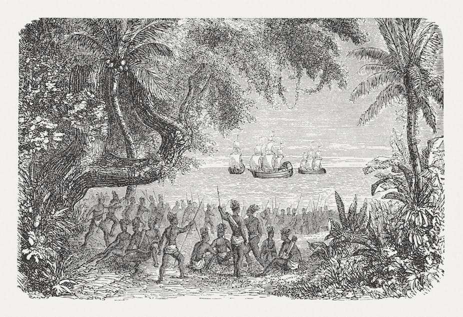 A black and white drawing shows a large crowd of Native American people wearing little clothing and holding spears, looking at boats in the water ahead of them.