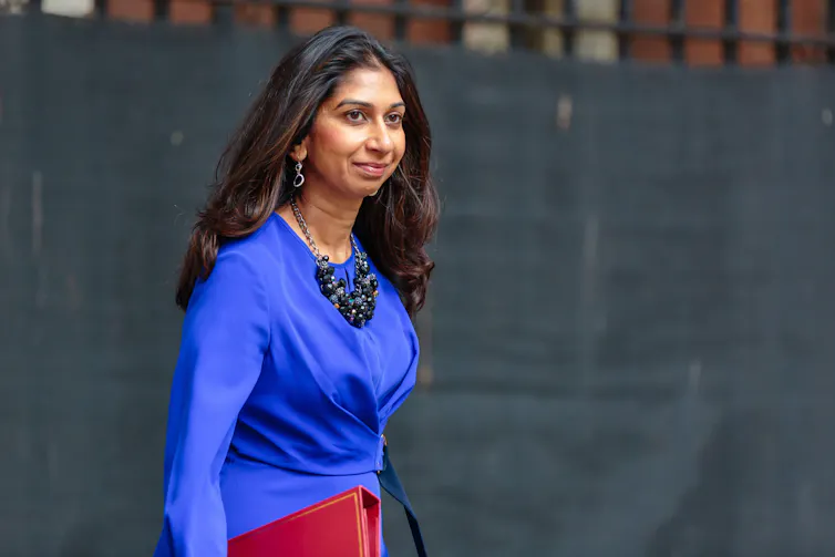 Suella Braverman standing outside in a blue dress and holding her red ministerial folder
