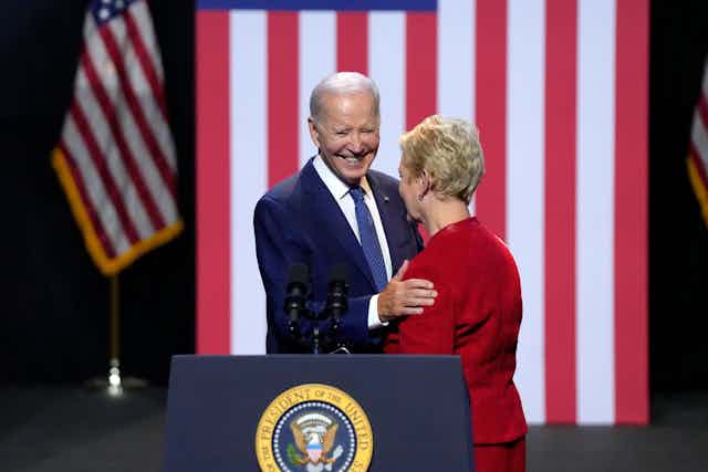 President Joe Biden talks to the widow of Republican senator John McCain at a speech he gave on democracy. He is standing at a podium with a flag behind him.