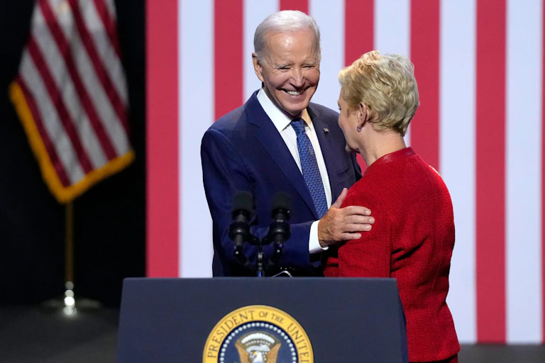 President Joe Biden talks to the widow of Republican senator John McCain at a speech he gave on democracy. He is standing at a podium with a flag behind him.