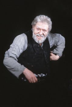 An old grey man in a grey shirt and black tank top on stage.