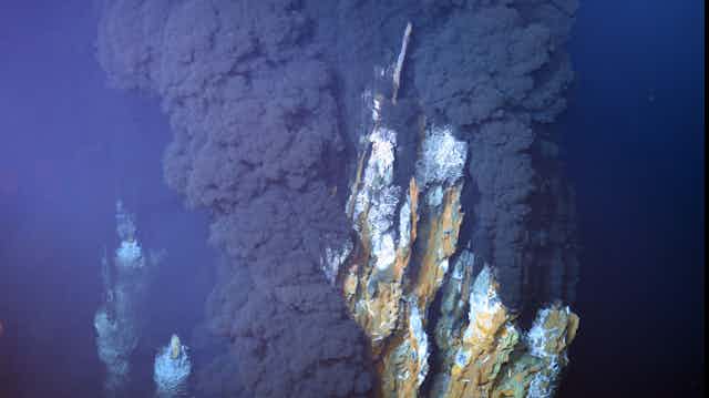 Cheminée hydrothermales marrons/grises/bleutées, avec un épais panache de fumée noire.