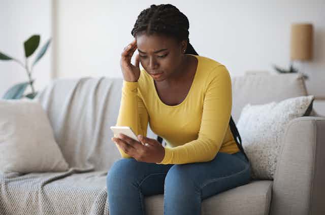 A young Black woman sits on a couch looking at a phone with a worried expression