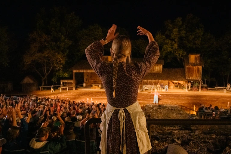 A woman with a braided ponytail raises her arms along with a large crowd of people at an outdoor performance.