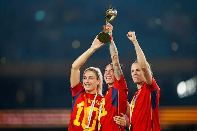 Three women in soccer jerseys hold up a trophy while wearing gold medals