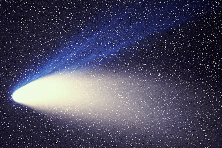 A bright comet streaking downward, with blue and white streaks of light following behind, against a backdrop of black and many small white stars.