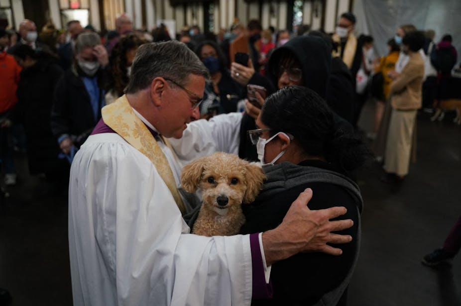 A man in a white clerical robe puts his hands on the shoulders of a woman in a white face mask who is holding a fuzzy dog.