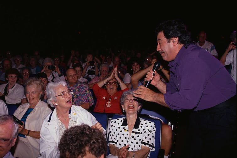 Man wearing purple shirt holding microphone singles out an elderly woman in the crowd.