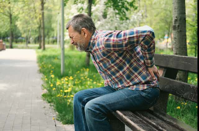 As he grimaces in pain, a man sitting on a park bench braces his back with his left hand.