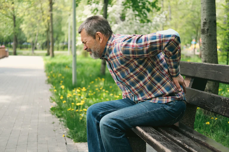 As he grimaces in pain, a man sitting on a park bench braces his back with his left hand.