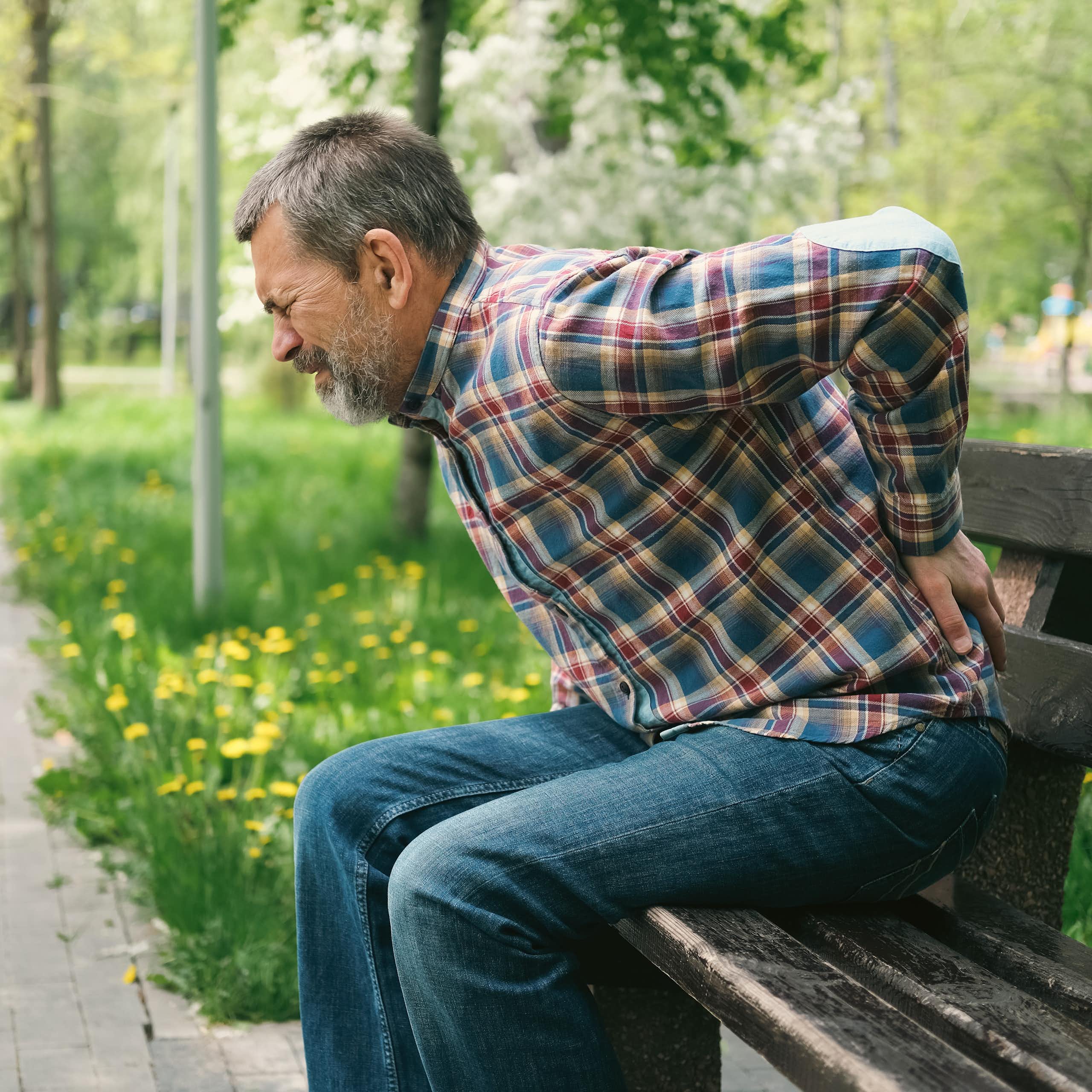 As he grimaces in pain, a man sitting on a park bench braces his back with his left hand.