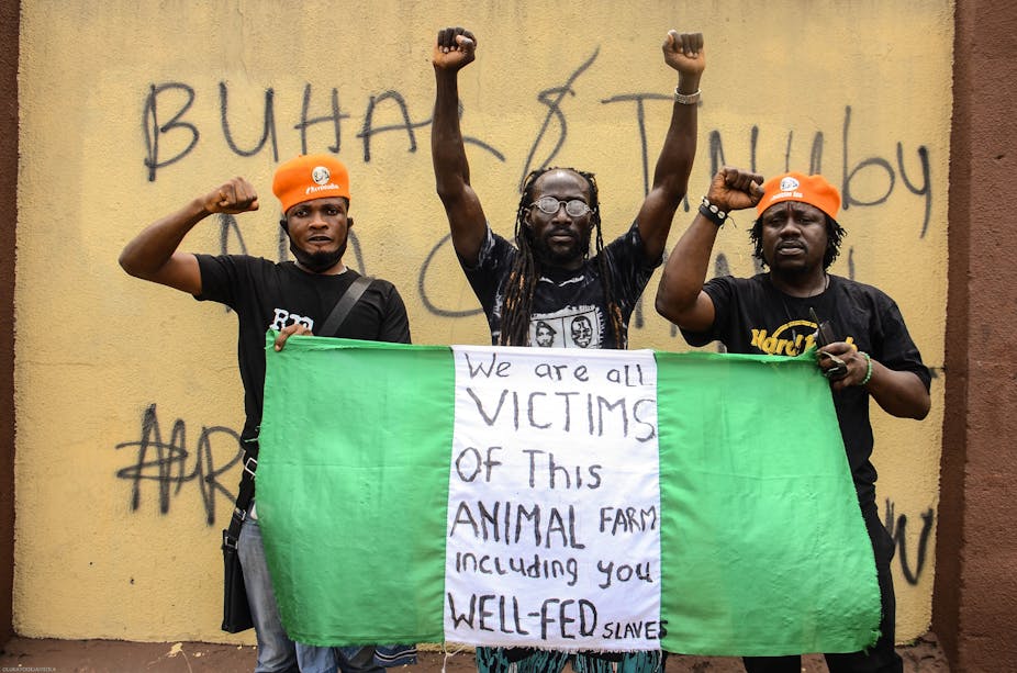 Three men holding a Nigerian flag during a protest. The flag reads "We are all victims of this animal farm including you well-fed slaves."