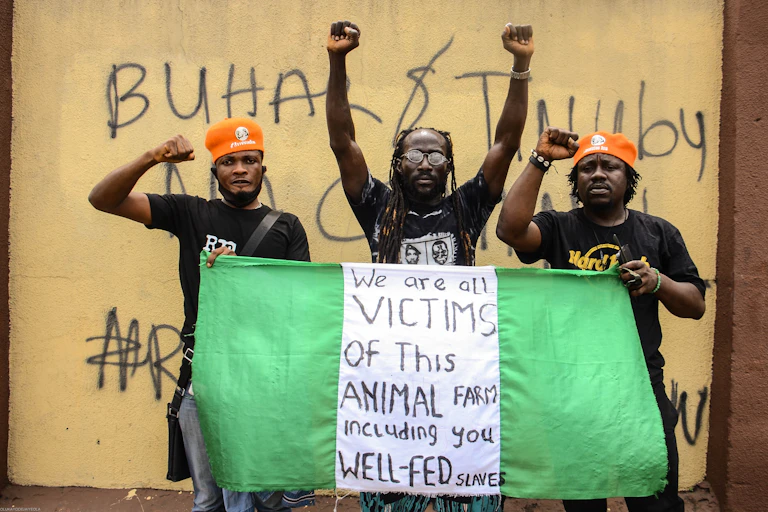Three men holding a Nigerian flag during a protest. The flag reads "We are all victims of this animal farm including you well-fed slaves."