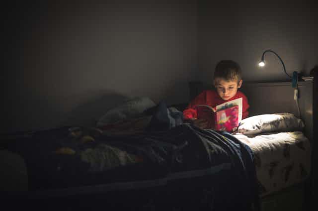 In a darkened room illuminated only by a reading lamp, a young boy in red pajamas sits upright in bed reading a book.