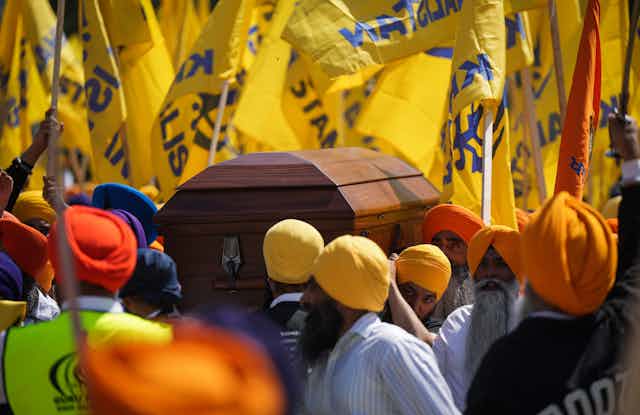 Men in orange and yellow turbans carry a casket. A sea of yellow flags is behind them.