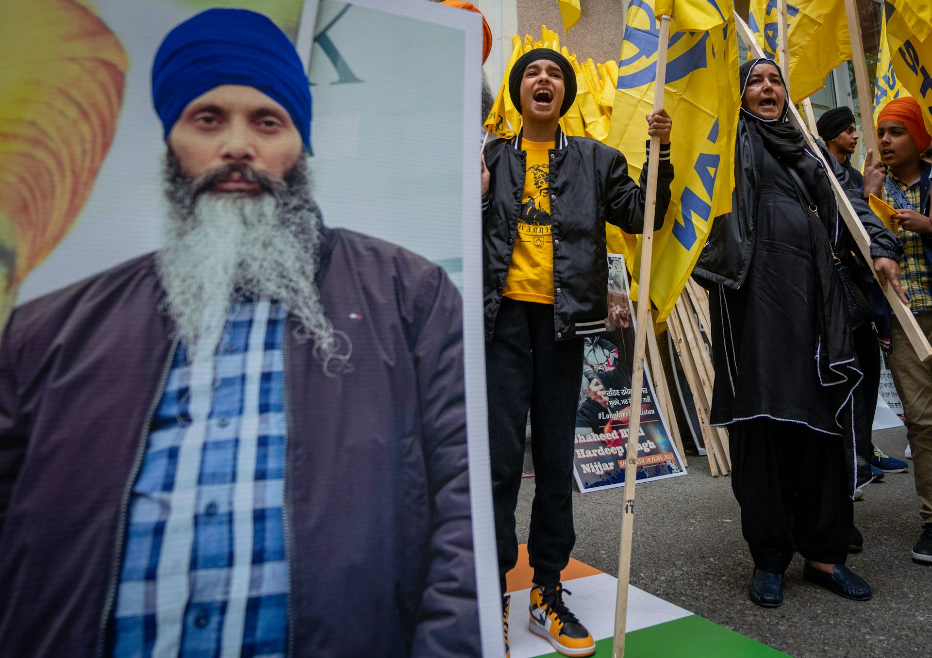 People hold signs and shout next to a large photo of a man with a grey beard and a blue turban.