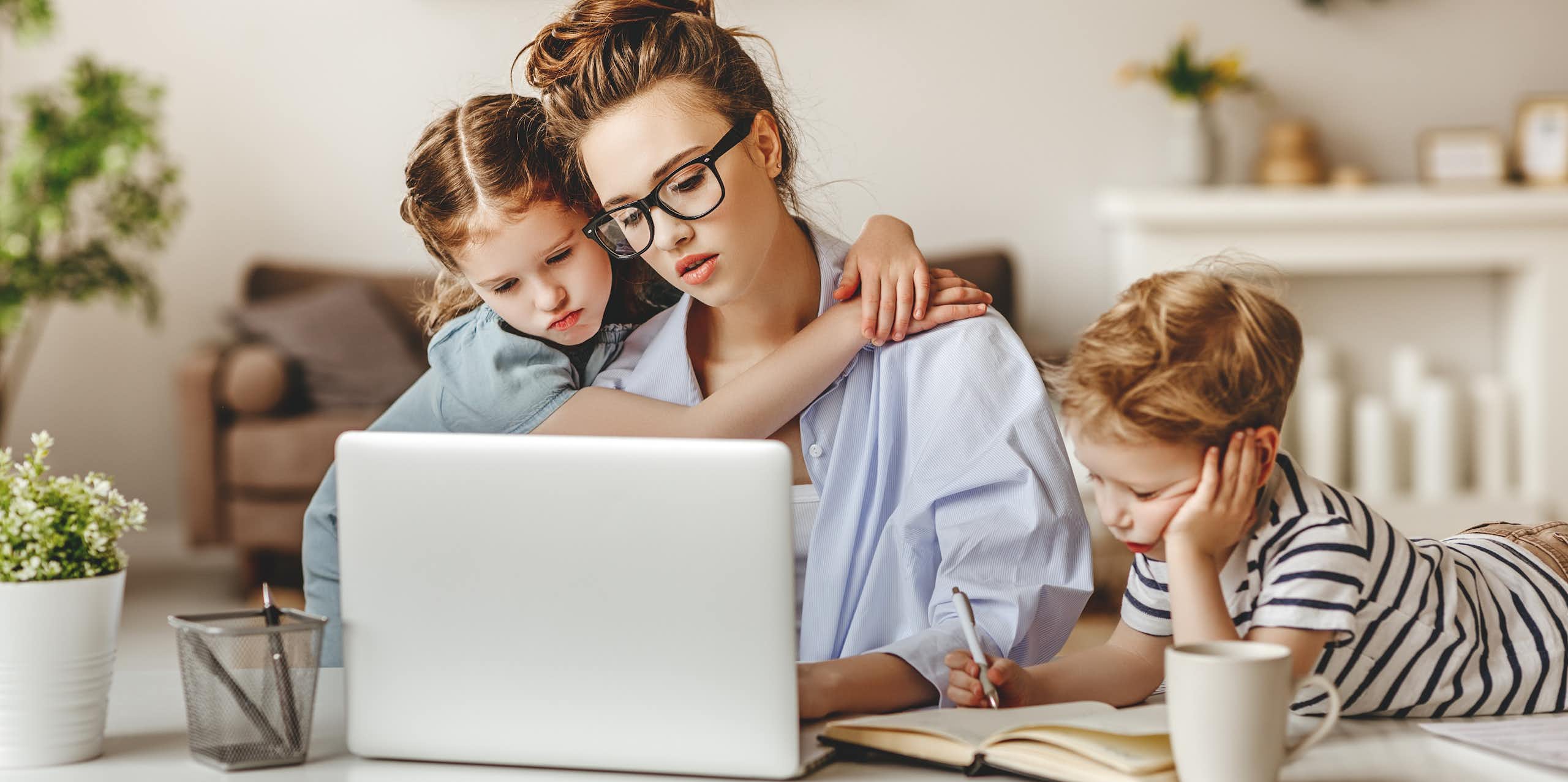 Una mujer joven escribe en el ordenador mientras una niña la abraza y un niño lee a su lado con cara de aburrido.
