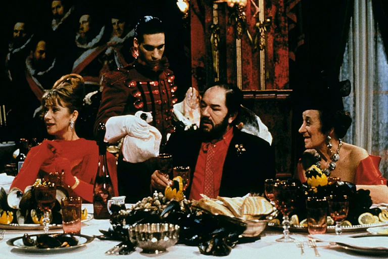 Albert Spica sits at a table with his wife and another woman.