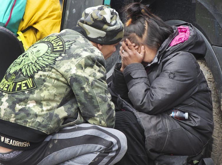 A woman cries with her face in her hands as someone crouches next to her.