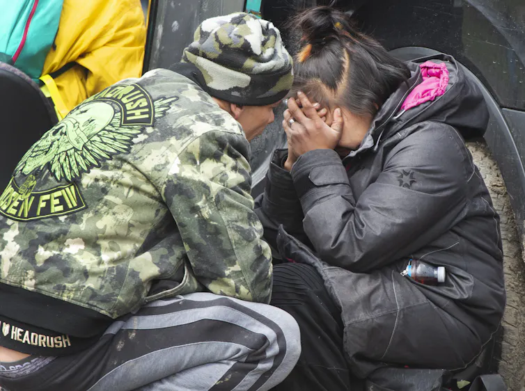 A woman cries with her face in her hands as someone crouches next to her.