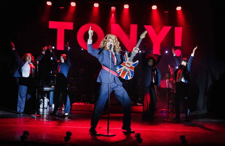 Man on stage in a long blonde wig holding a union jack guitar.