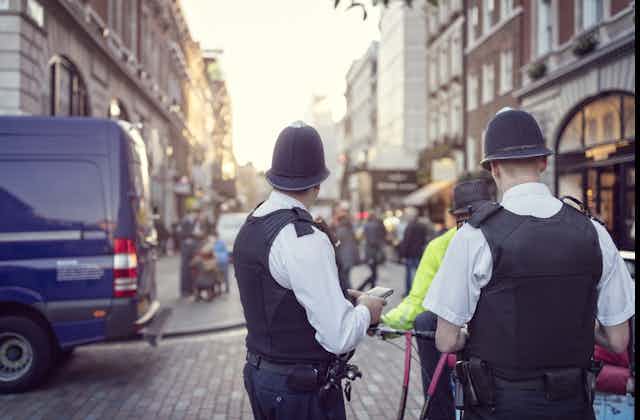 Two police officers from the back look down a high street with shops.