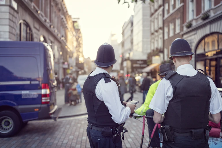 Two police officers from the back look down a high street with shops.