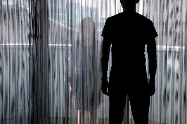 Silhouette of woman looking through a curtain at a shadowed man in the foreground.