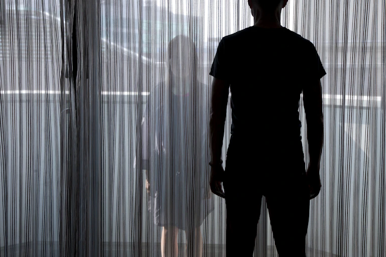 Silhouette of woman looking through a curtain at a shadowed man in the foreground.