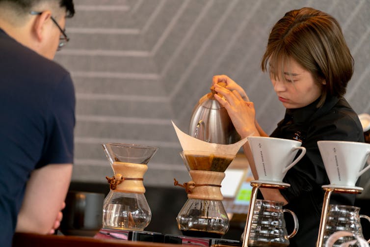 A young female Chinese barista pours hot water as a male customer awaits his order.