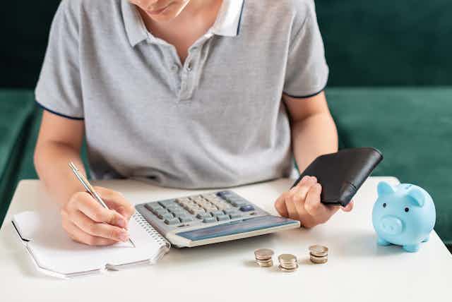 Un adolescente lleva cuentas con calculadora y una libreta mientras tiene montoncitos de monedas en la mesa.