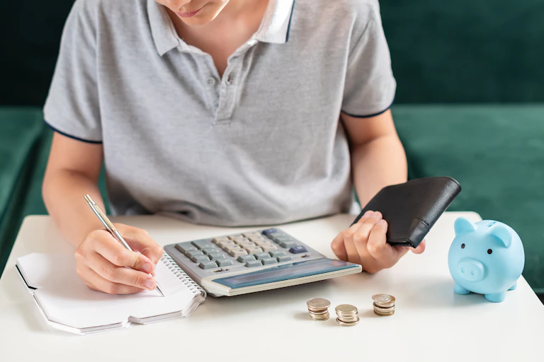 Un adolescente lleva cuentas con calculadora y una libreta mientras tiene montoncitos de monedas en la mesa.