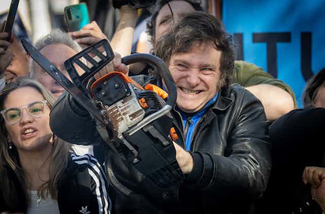 A man with a grin on his face carries a chain saw while surrounded by onlookers.
