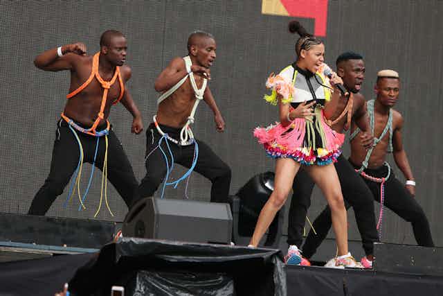 A young woman sings into a microphone on stage, backed by four males dancers bare chested, wearing beads. She wears a multi-layered short bright pink skirt, a midriff blouse with traditional African trimmings and her hair is cornrowed with a large pointed hairpiece.