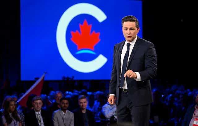 Un homme en cravate prend la parole devant une assemblée, avec un logo affiché derrière lui